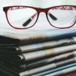 Red eyeglasses resting on a stack of newspapers symbolize reading and information.